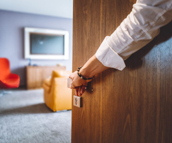 Toned man closing the door of the luxurious hotel room, man wearing a bracelet and a white shirt, closing the wooden door, nice and luxurious hotel room in the background.